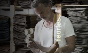 a man staring down at leather, he has a measuring tape around his neck. There are other strips of fabric and leather stacked up in the back on shelves. The word fealess is over the image.
