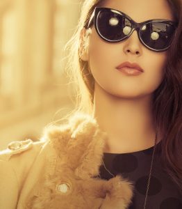 Women in sunglasses with hair to the left side. She has lip stick on and a coat with fur access.