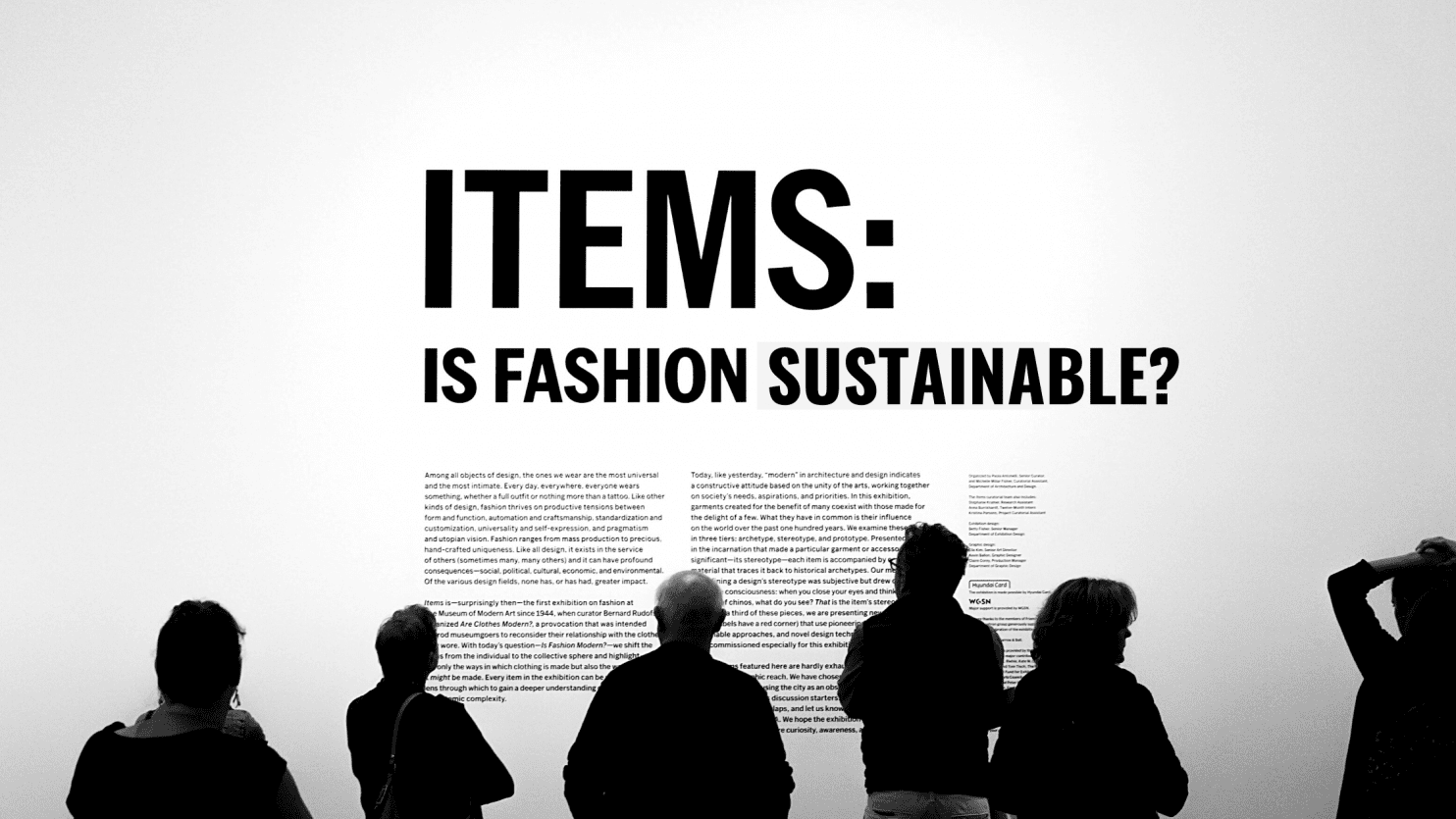 Black and white image of people looking at a art exhibit description that says "Items: Is Fashion Sustainable?"