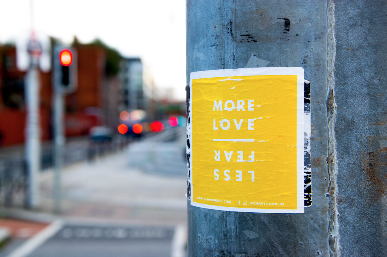 A sticker on a pole in a city that is yellow and says "more love" and than "less fear" upside down below it.