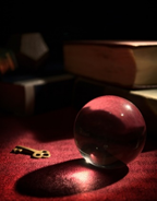 Background are books but are hard to see due to the dark lighting of the photo. There is a glass ball and key in the front of a red table.