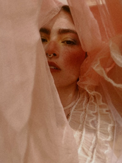 Editorial image of a women with a white sheer fabric half over her face. The color a peach hue.