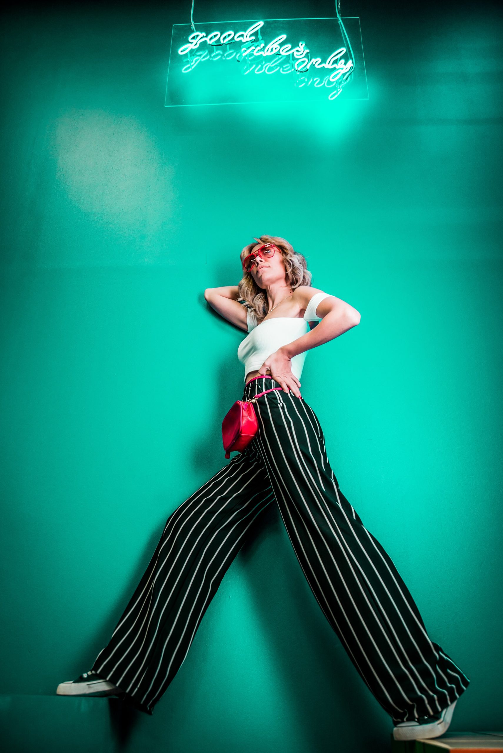 Editoral photo of a women in black and white stripped pants and a white shirt. The background is a seafoam green with a neon sign that says "good vibes only"