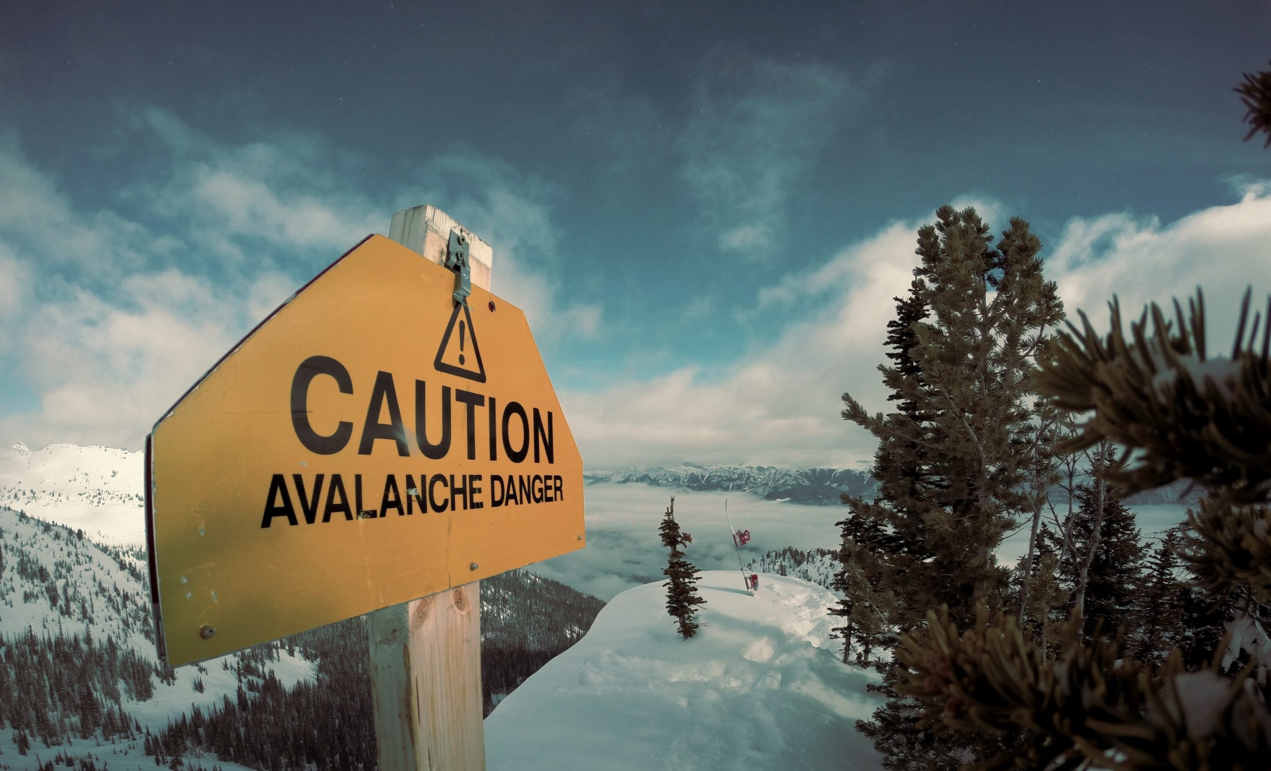 Sign that says "Caution Avalanche Danger", background is winter mountain view.