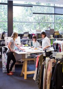 Three women at a table that is laid out with apparel designs with clothing and fashion books all around them